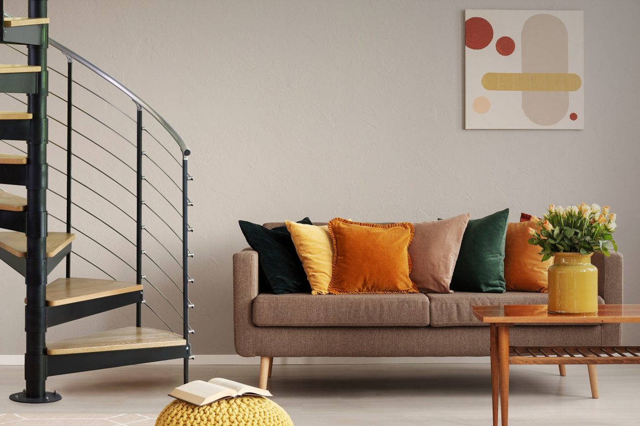 modern living room with beige sofa and yellow decor next to a black spiral staircase with silver gray multine railing and light wood treads